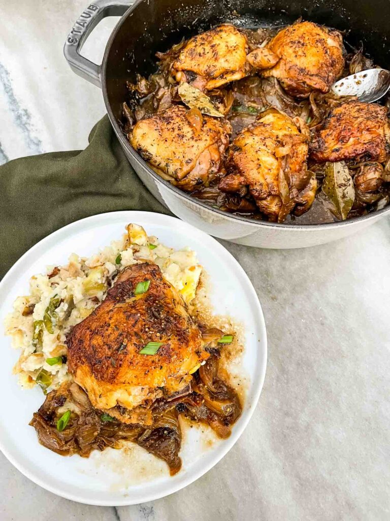 Top view of one piece of chicken on a white plate next to the braiser full of chicken on a counter