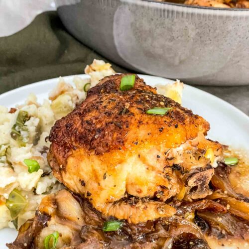 A chicken thigh sitting on colcannon on a white plate with the sauce and onions under it