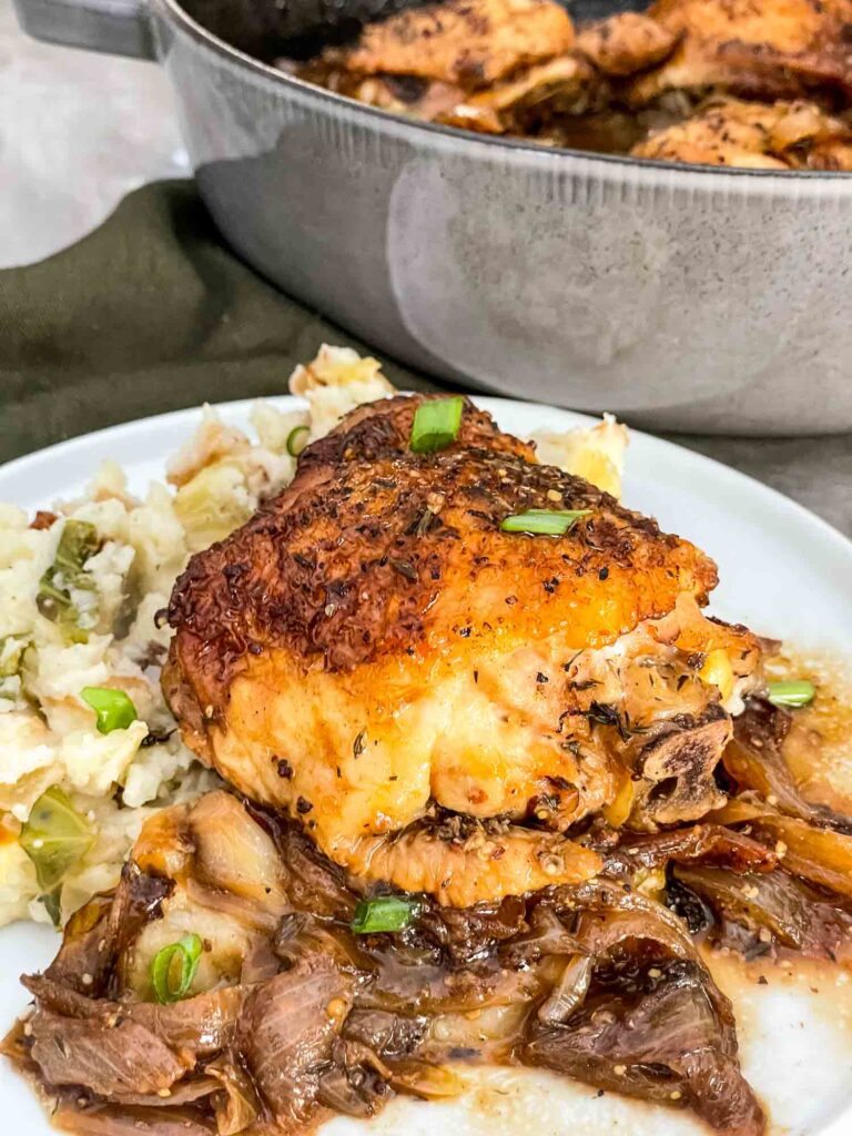 A chicken thigh sitting on colcannon on a white plate with the sauce and onions under it