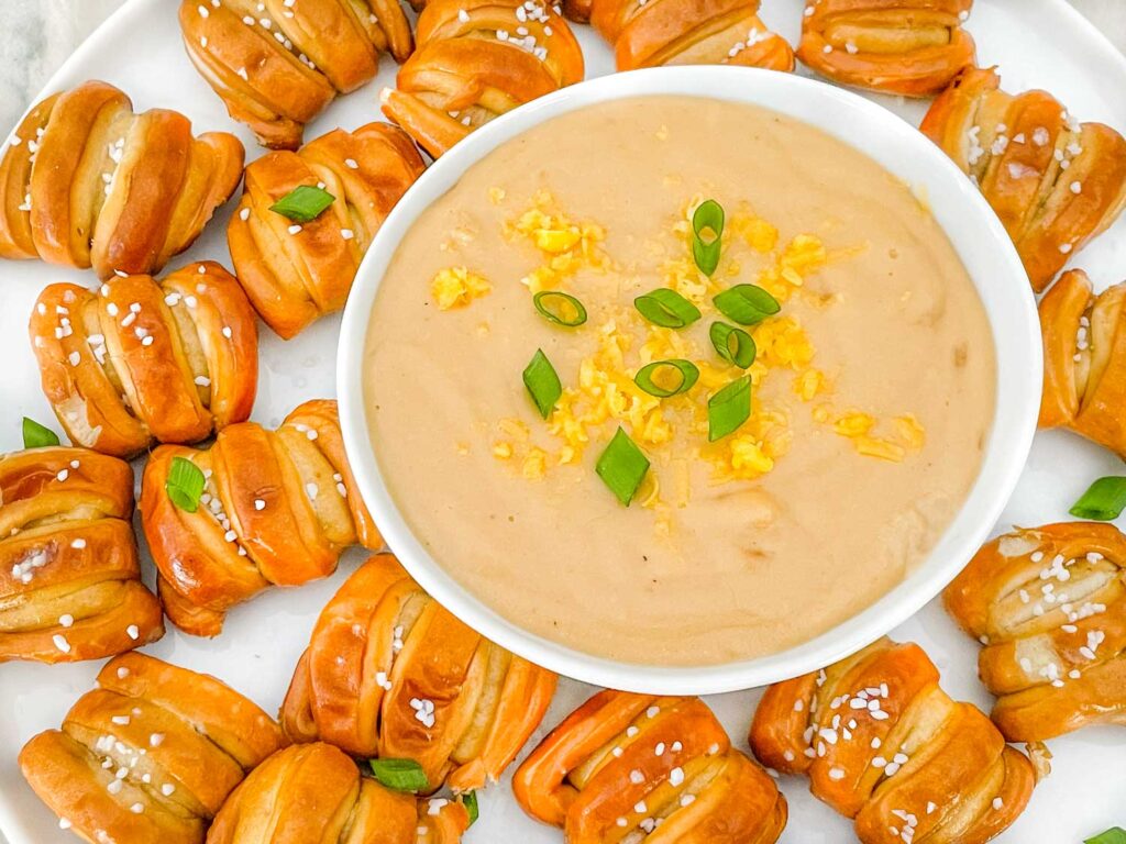A bowl of Beer Cheese Dip sitting on a plate full of soft pretzel pieces