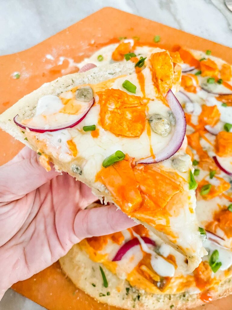 A hand holding a slice of Buffalo Chicken Pizza