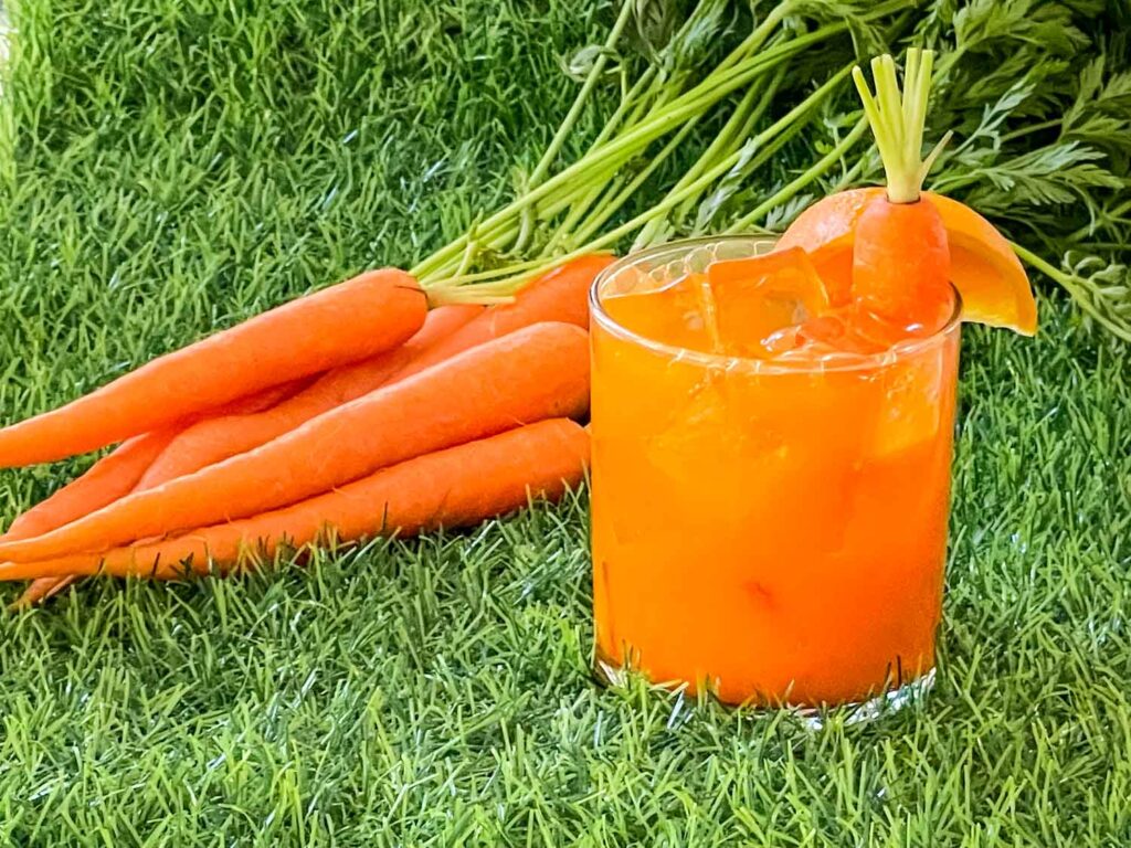 An orange Carrot Patch Mocktail in a short glass sitting on grass with a bunch of carrots behind it