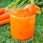 A glass of Carrot Patch Mocktail sitting on grass with some carrots behind it