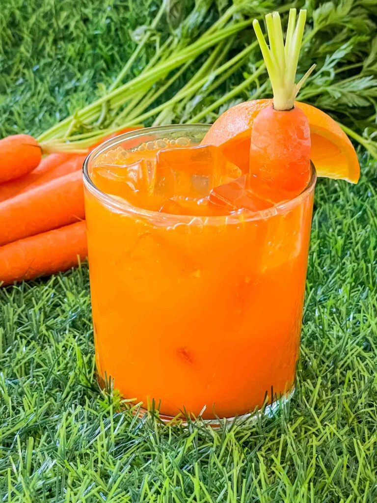 A glass of Carrot Patch Mocktail sitting on grass with some carrots behind it