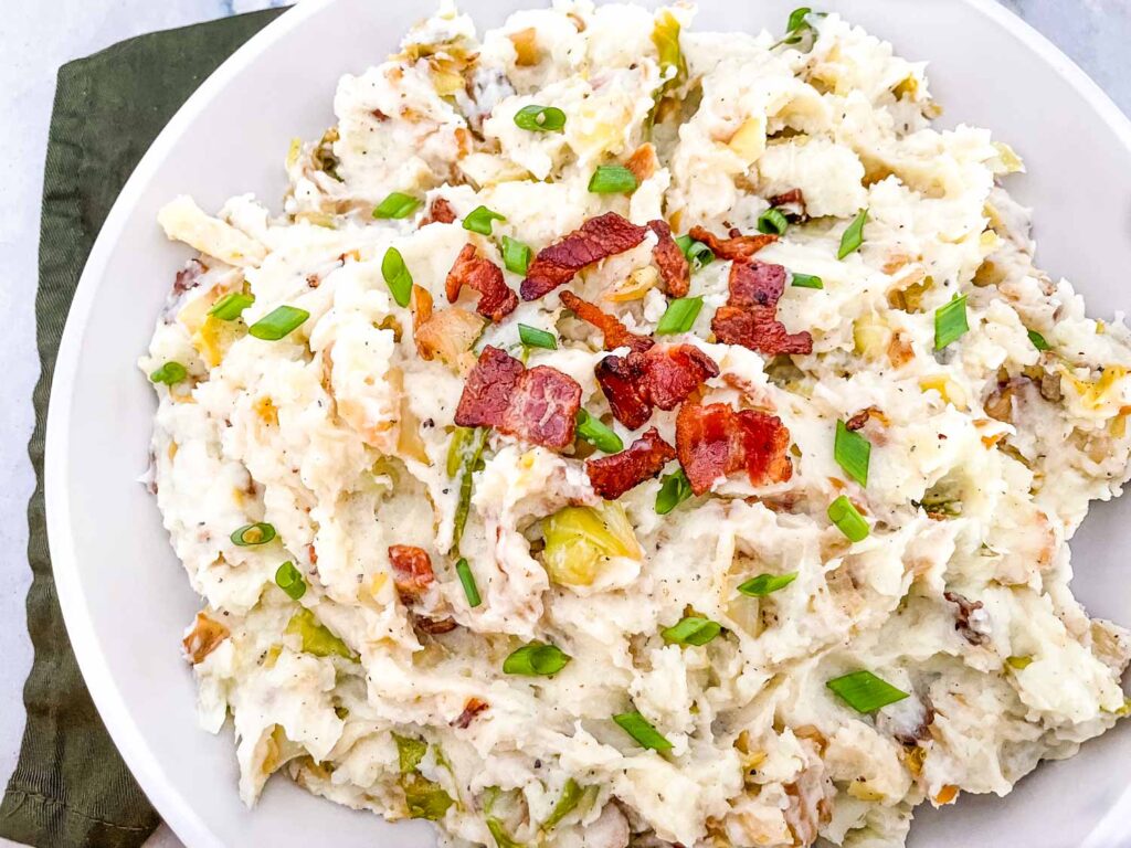 Top view of Colcannon with Bacon in a serving dish sitting on a green napkin on a counter. Garnished with more bacon and green onion pieces