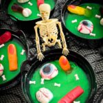 Pin of a skeleton standing at a Halloween Jello Cauldron with gummy body parts in it and title at top