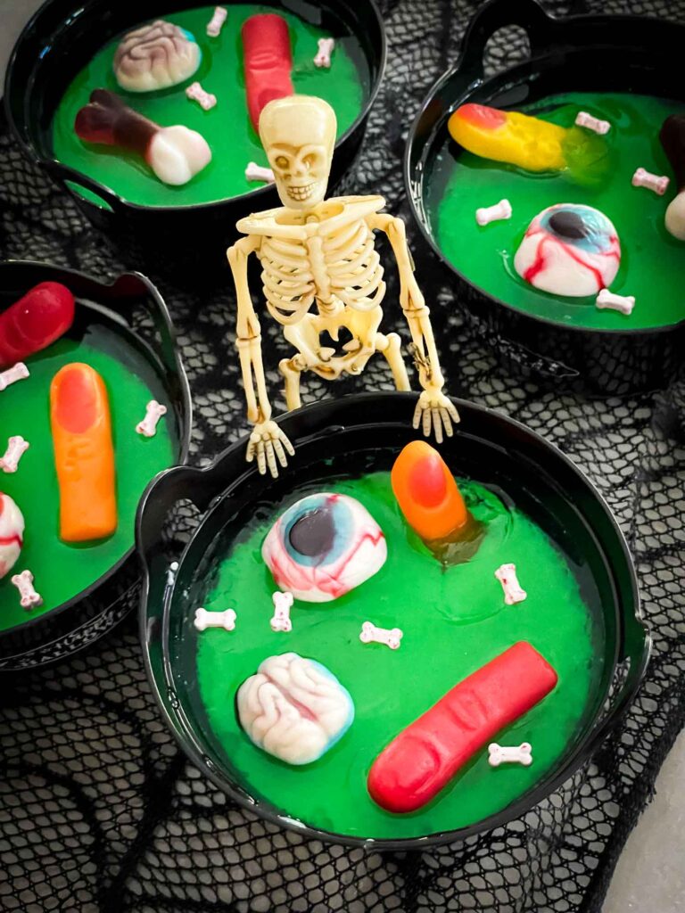 A skeleton standing at a green Halloween Jello Cauldron with gummy body pieces in it