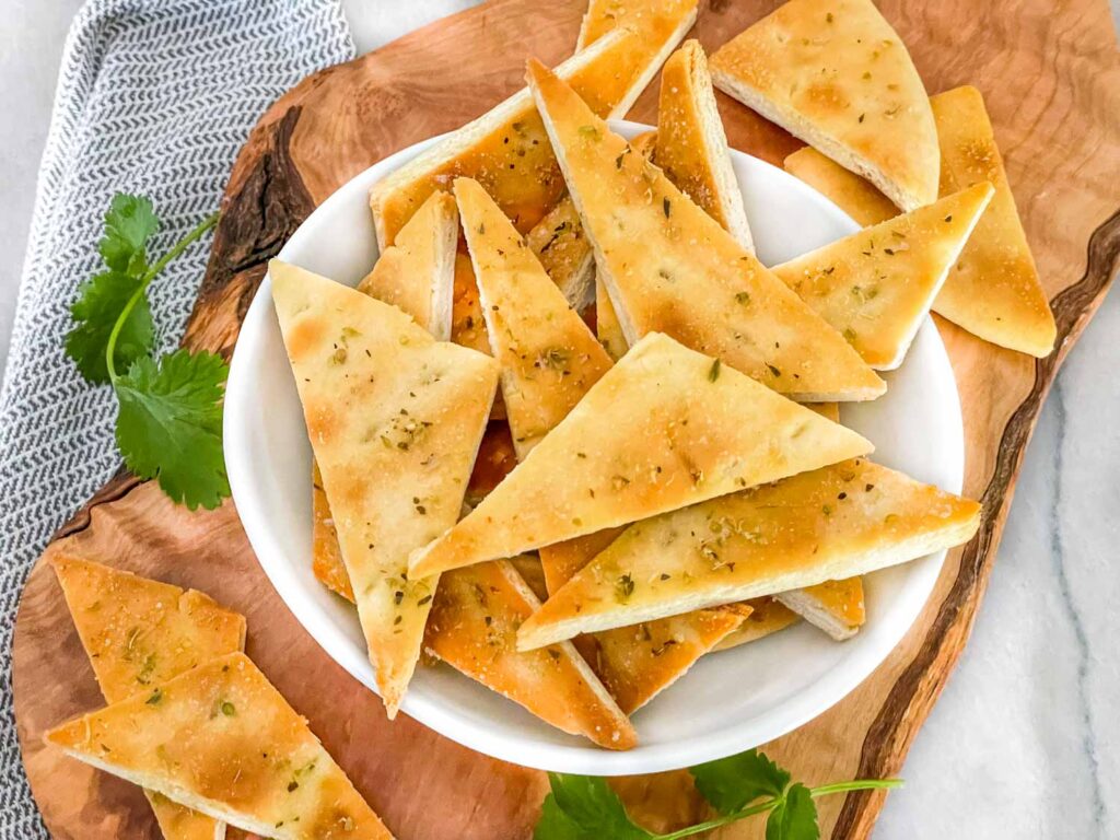 Homemade Baked Pita Chips piled in a small white bowl sitting on a wood board on a counter with chips around it