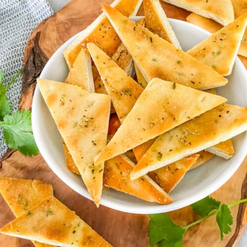 Small Homemade Baked Pita Chips triangles piled in a small white bowl on a wood board sitting on a counter