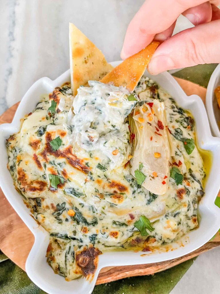 A hand dunking pita chips into the Easy Spinach Artichoke Dip