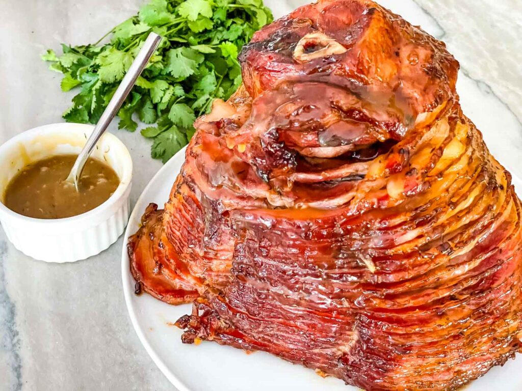 The entire ham on a white plate with a bowl of the extra glaze next to it on a counter