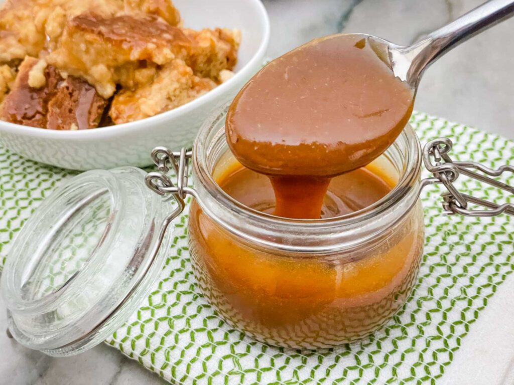 Irish Cream Dessert Sauce in a clear glass jar with a spoon coming out of the sauce. Sitting on a green and white towel in front of bread pudding