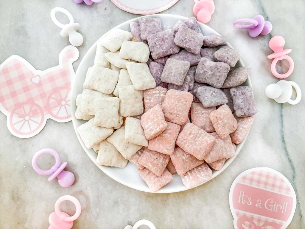 A small white plate with purple, pink, and white It's a Girl! Puppy Chow on it sitting on a counter with decorations