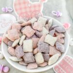 A small white bowl full of purple, pink, and white It's a Girl! Puppy Chow on a counter with baby decor