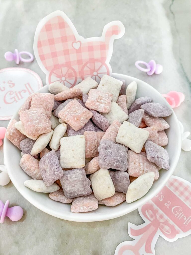 A small white bowl full of purple, pink, and white It's a Girl! Puppy Chow on a counter with baby decor