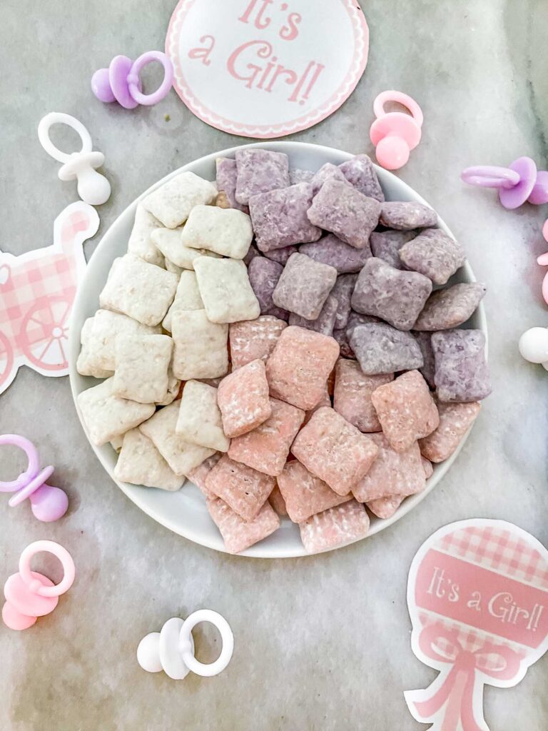 Purple, pink, and white muddy buddies on a small plate on a counter with baby shower decorations around it