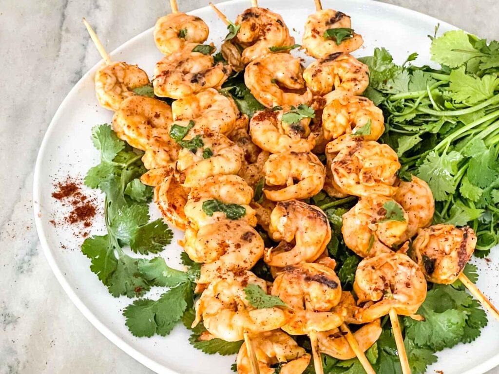 Mexican Grilled Shrimp on skewers sitting on cilantro on a white plate on a counter