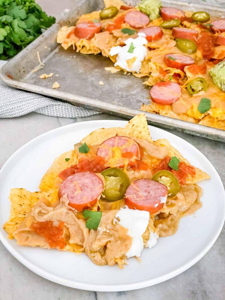 Some Sausage Beer Cheese Nachos on a white plate with a baking sheet of nachos behind it