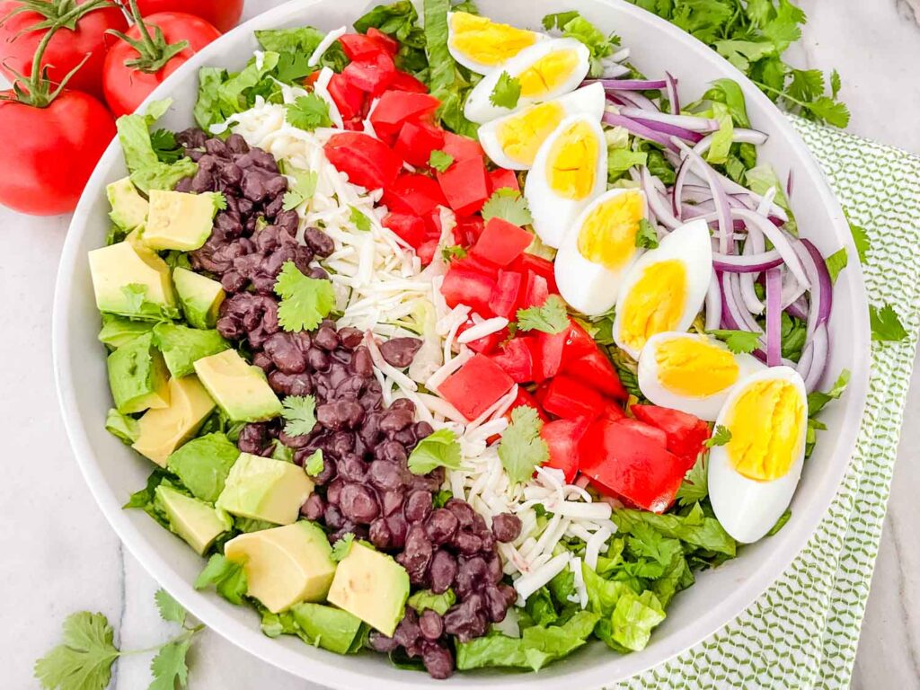 Top view of Mexican Cobb Salad with avocado, black beans, tomato, cheese, egg, and onion on top of lettuce in rows