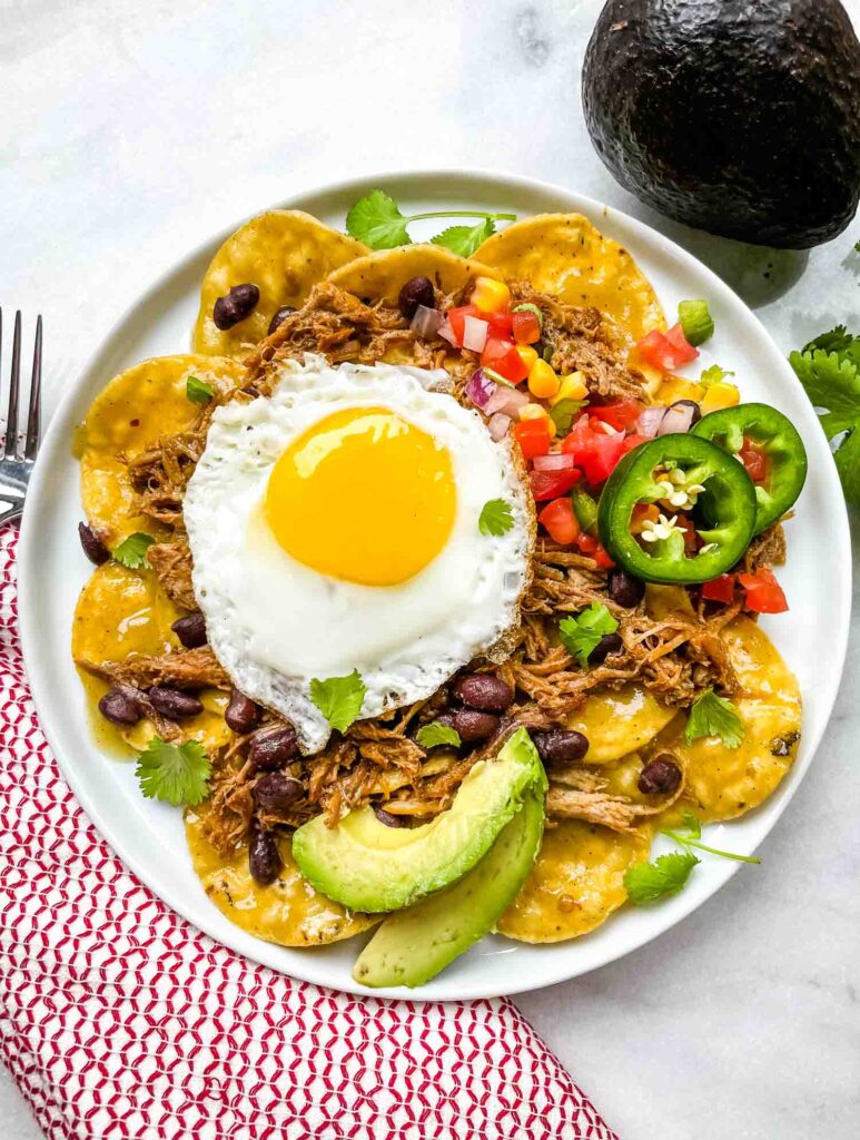 Top view of a plate of Pork Carnitas Green Chilaquiles with Eggs with a red towel and avocado next to it