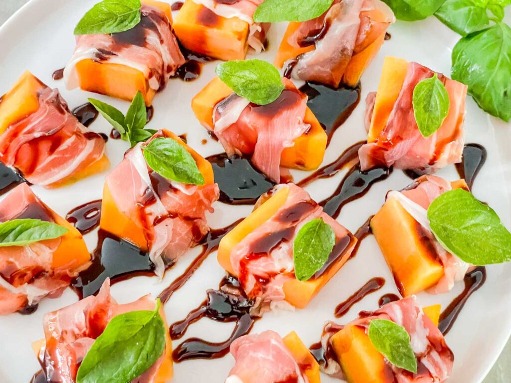 A white plate filled with Prosciutto Wrapped Melon with Balsamic Glaze drizzled across them all