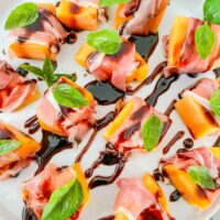 A white plate full of Prosciutto Wrapped Melon with Balsamic Glaze pieces