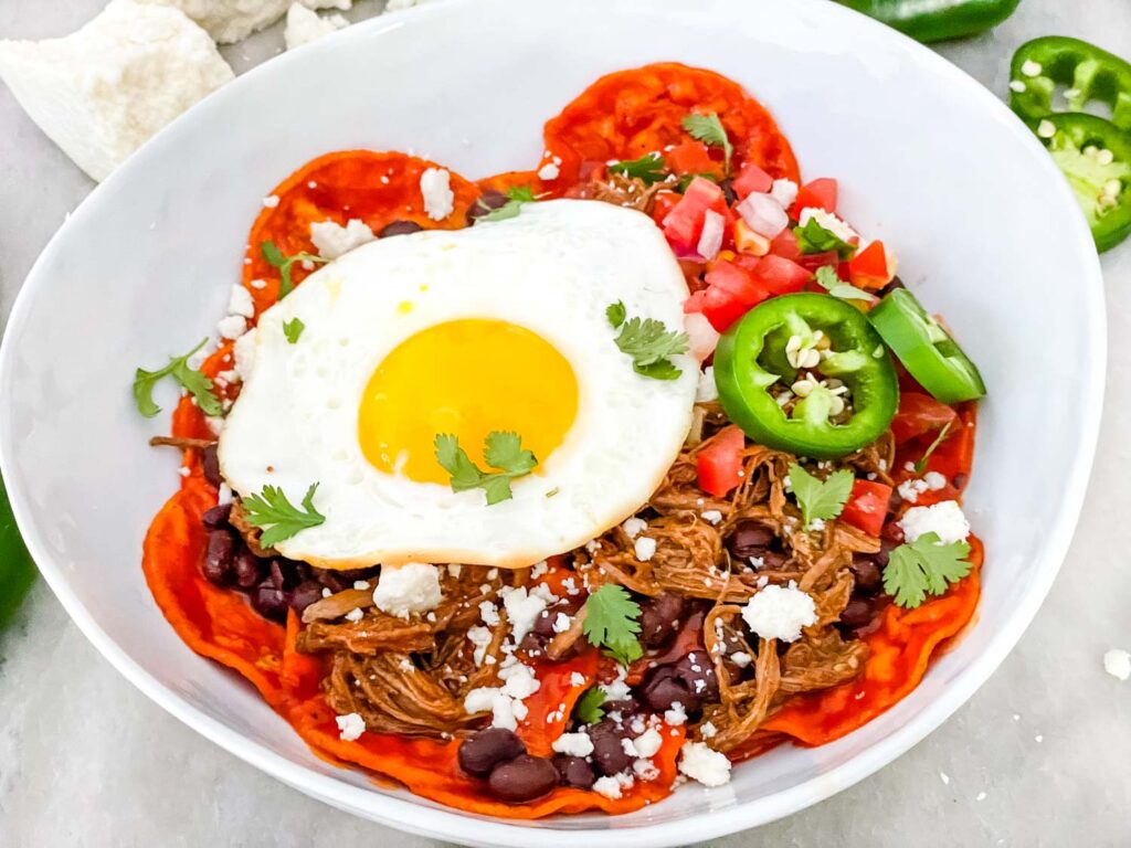 Shredded Beef Red Chilaquiles in a white bowl with an egg on top. Sitting on a counter