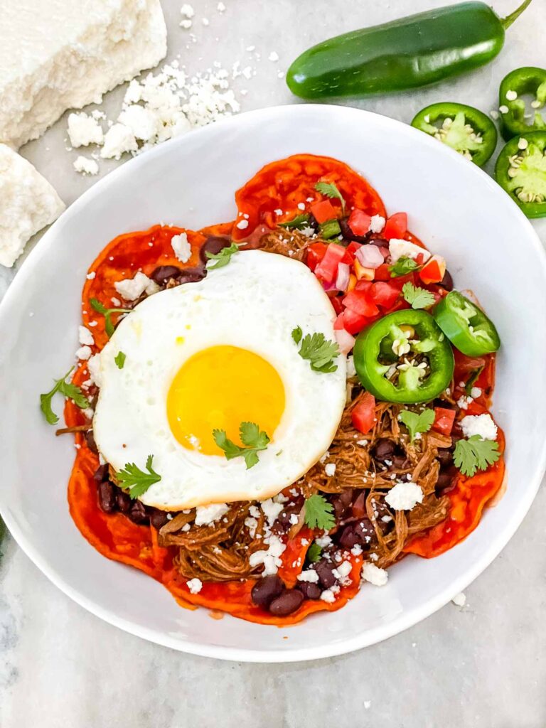 Shredded Beef Red Chilaquiles with an egg on top in a bowl from the top view sitting on a counter surrounded by peppers and cheese