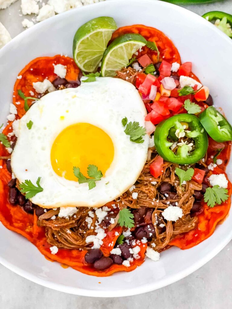 Top view of Shredded Beef Red Chilaquiles in a white bowl with an egg and limes