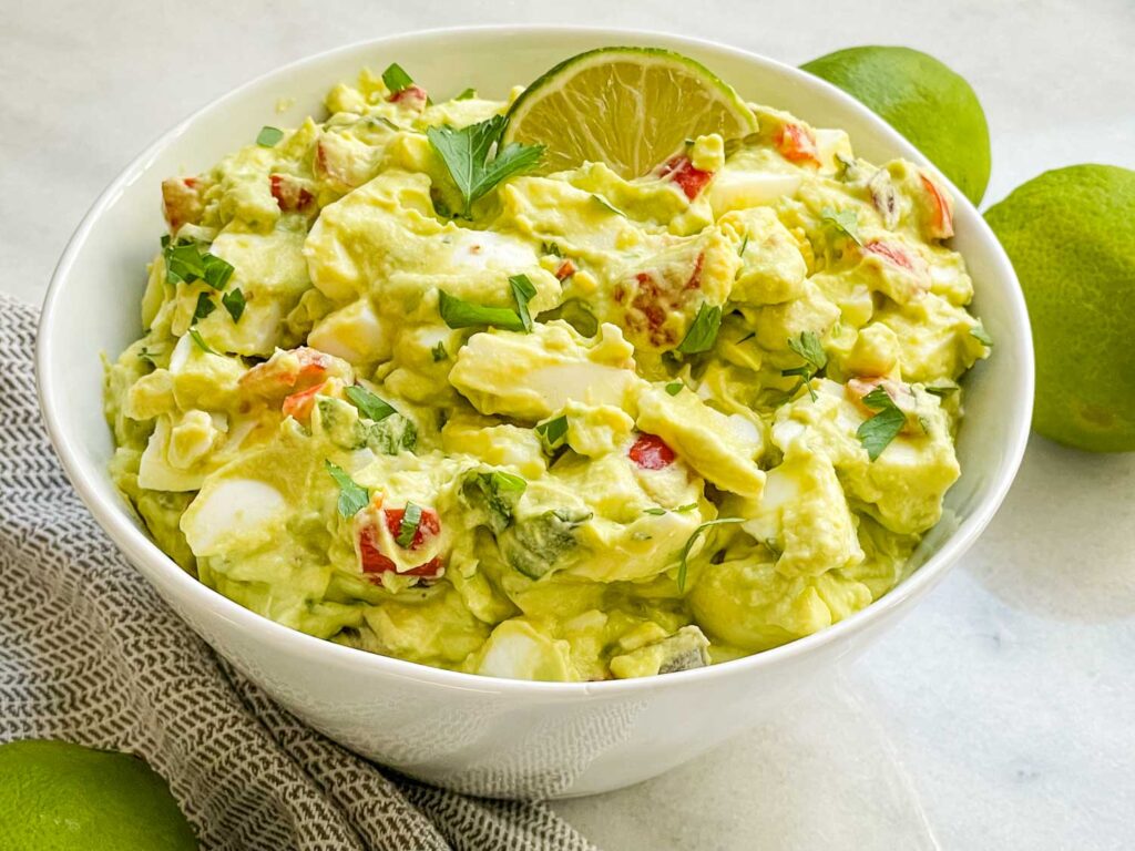 A white bowl full of Avocado Egg Salad on a counter with limes and a towel