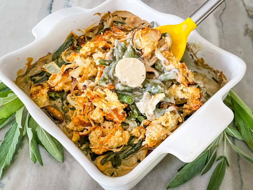 A spoon scooping some Green Bean Casserole with Fresh Green Beans out of the dish on a counter with fresh sage around it