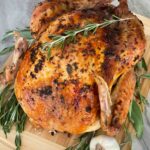 A whole Italian Herb Butter Roasted Turkey on a cutting board with fresh herbs and garlic next to it