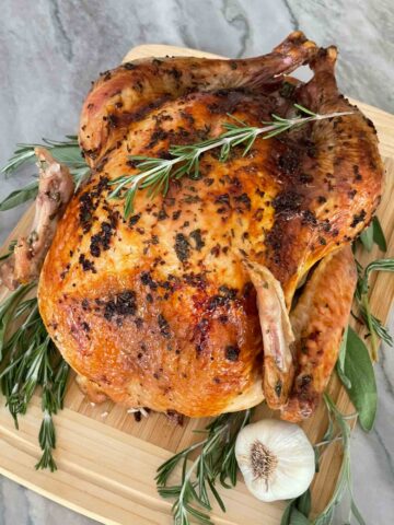 A whole Italian Herb Butter Roasted Turkey on a cutting board with fresh herbs and garlic next to it