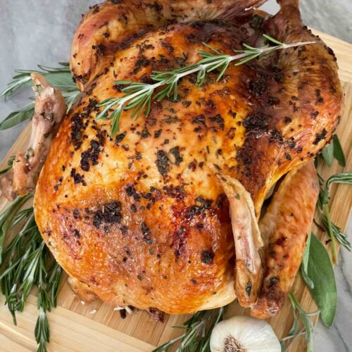 A whole Italian Herb Butter Roasted Turkey on a cutting board with fresh herbs and garlic next to it