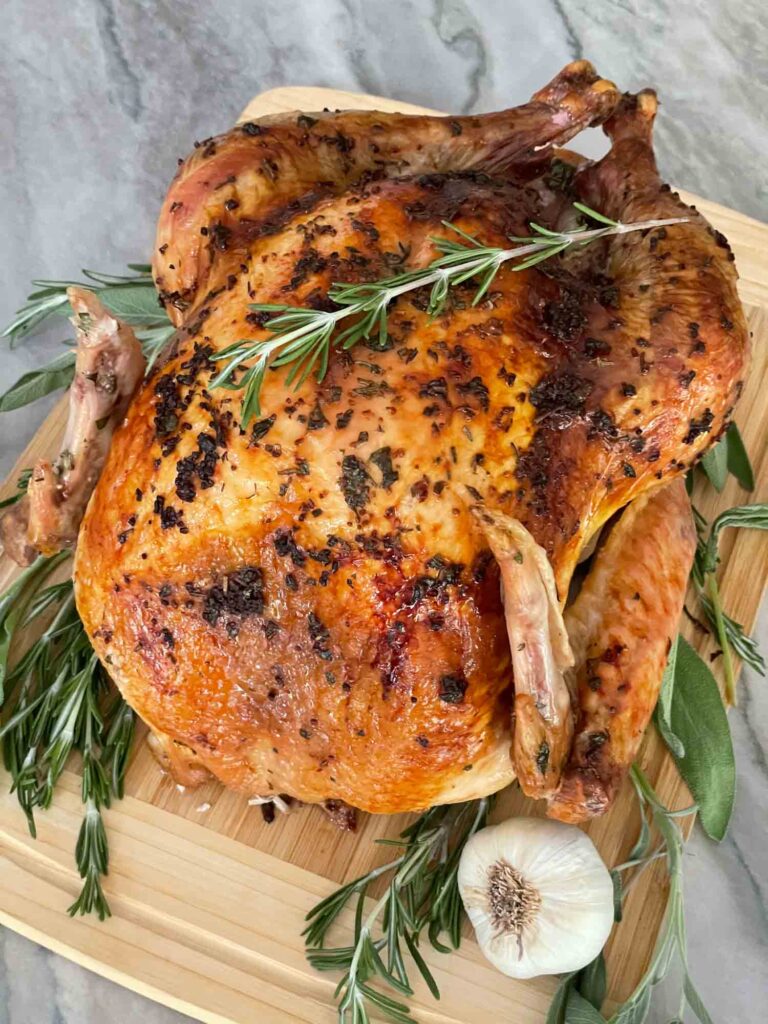A whole Italian Herb Butter Roasted Turkey on a cutting board with fresh herbs and garlic next to it