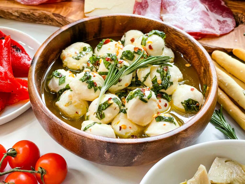 A wood bowl full of Marinated Mozzarella Balls surrounded by charcuterie ingredients