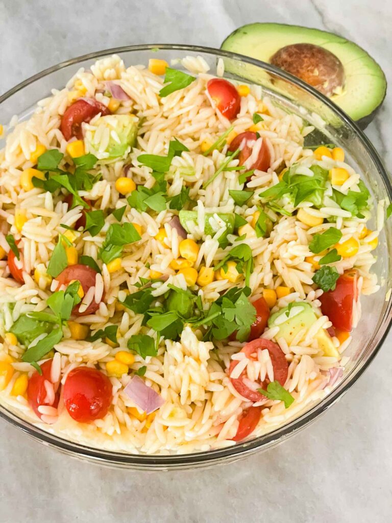 Top view of Mexican Orzo Pasta Salad in a glass bowl on a counter next to an avocado