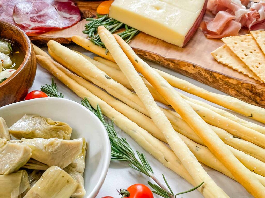 A pile of Rosemary Grissini breadsticks on a counter surrounded by Italian ingredients