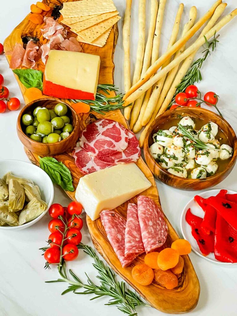 Top view of a Antipasti Italian Charcuterie Board on a white counter. Thin breadsticks, meats, cheeses, olives, and other foods.