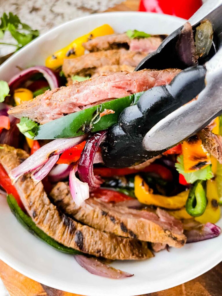 Tongs picking up some Chipotle Steak Fajitas with peppers and onions