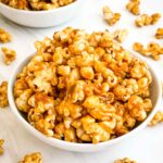 A white bowl full of Homemade Caramel Popcorn Recipe (without Corn Syrup) on a white counter.