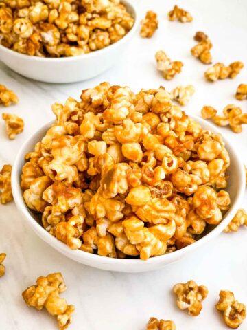 A white bowl full of Homemade Caramel Popcorn Recipe (without Corn Syrup) on a white counter.