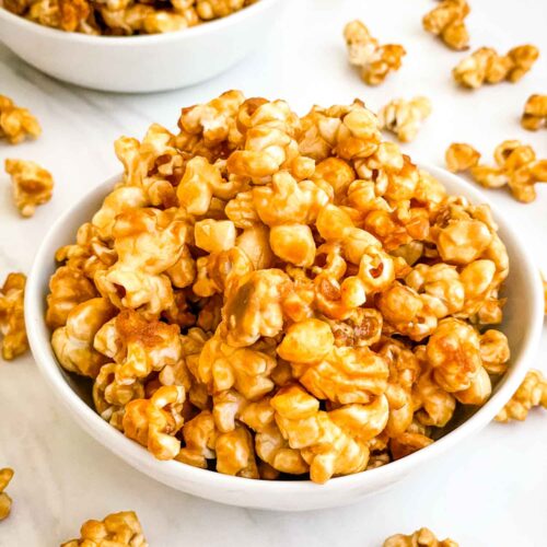 A white bowl full of Homemade Caramel Popcorn Recipe (without Corn Syrup) on a white counter.