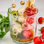 One Individual Italian Charcuterie Cup on a counter with basil and tomatoes around it.