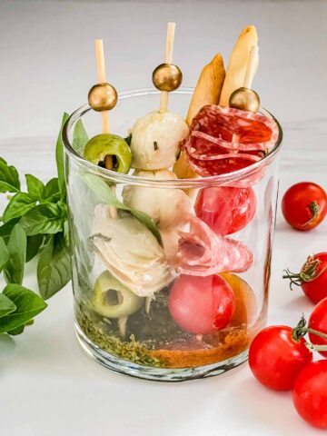 One Individual Italian Charcuterie Cup on a counter with basil and tomatoes around it.