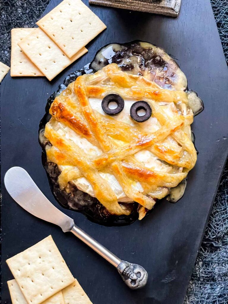 Looking down on a Halloween Mummy Wrapped Baked Brie Recipe on a black board.