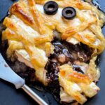 A Halloween Mummy Wrapped Baked Brie Recipe cut open on a black board with cheese and red jam coming out of it.
