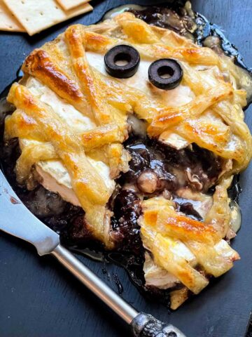 A Halloween Mummy Wrapped Baked Brie Recipe cut open on a black board with cheese and red jam coming out of it.