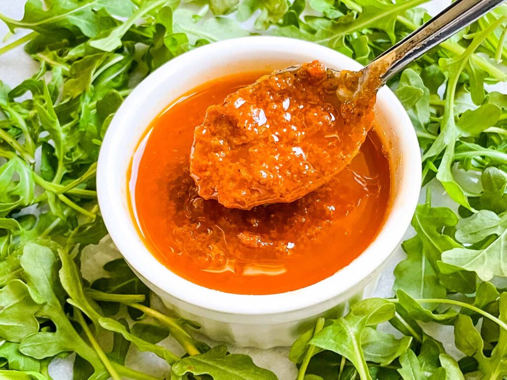 Sundried Tomato Vinaigrette Salad Dressing in a small white dish with a spoon scooping some out, surrounded by arugula.