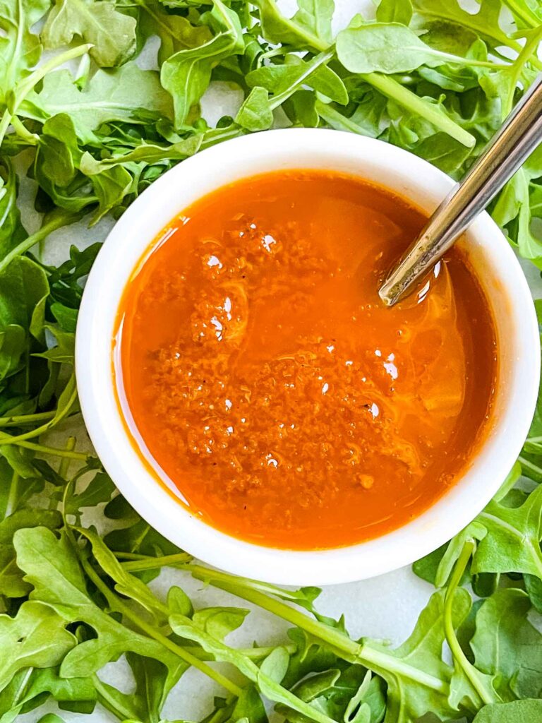 Looking down on a small white dish full of Sundried Tomato Vinaigrette Salad Dressing surrounded by arugula lettuce.